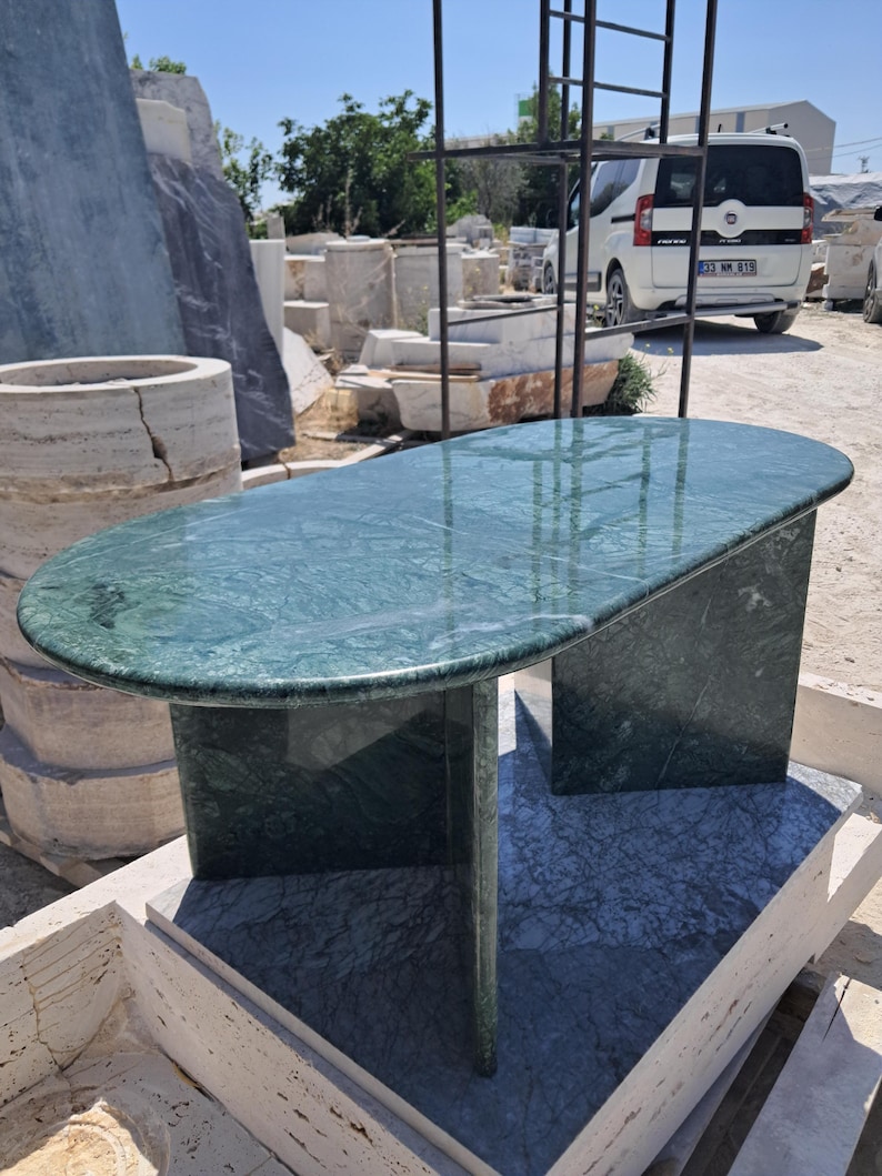 Puede incluir: Una mesa de centro de m&aacute;rmol verde con una tapa rectangular redondeada y dos patas de pedestal. La mesa est&aacute; hecha de m&aacute;rmol pulido con vetas naturales. La mesa est&aacute; al aire libre en un patio de piedra.