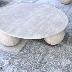 May include: A round, light-colored marble coffee table with a textured surface. The table rests on three rounded, ball-shaped legs. The table is set on a stone patio.