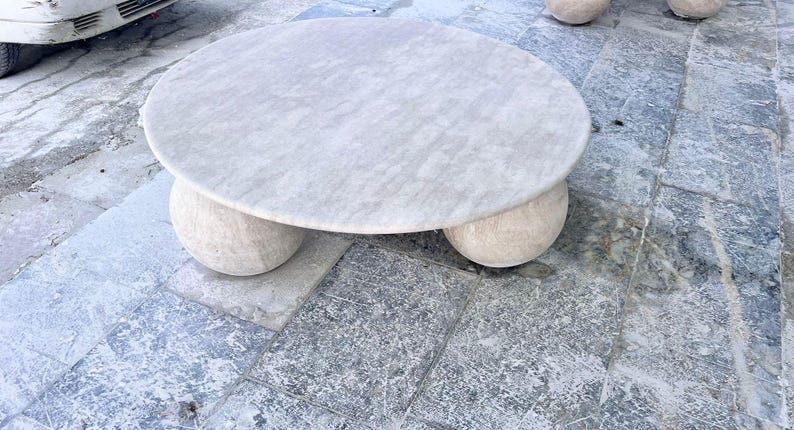 May include: A round, light-colored marble coffee table with a textured surface. The table rests on three rounded, ball-shaped legs. The table is set on a stone patio.