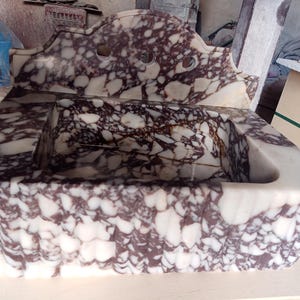 May include: A rectangular marble sink with a decorative backsplash. The sink features a white and burgundy patterned design. The backsplash has three circular cutouts. The sink is made of stone and has a carved, textured exterior.