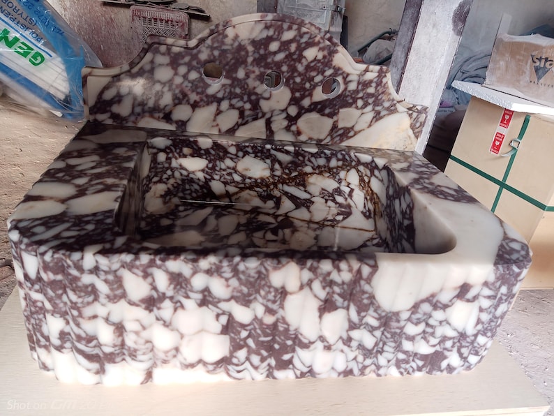 May include: A rectangular marble sink with a decorative backsplash. The sink features a white and burgundy patterned design. The backsplash has three circular cutouts. The sink is made of stone and has a carved, textured exterior.