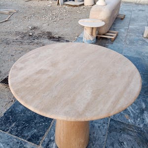 May include: A round, beige marble table with a cylindrical base. The table has a smooth, polished surface and is set outdoors. The table is approximately 40 inches in diameter.