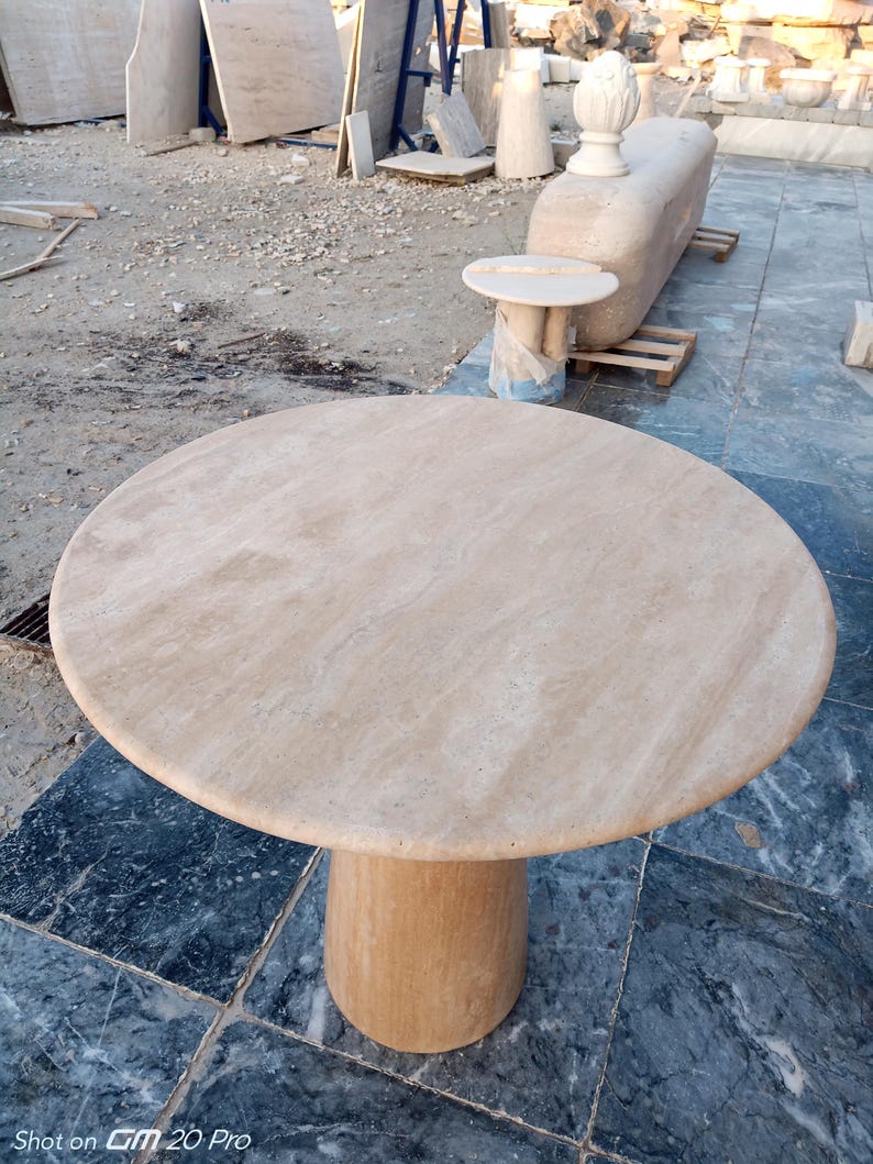 May include: A round, beige marble table with a cylindrical base. The table has a smooth, polished surface and is set outdoors. The table is approximately 40 inches in diameter.