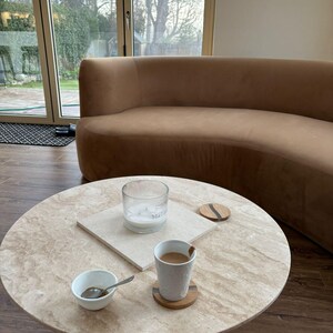 May include: A round, light-colored stone coffee table with a pedestal base. A small white bowl with a spoon, a cup of coffee, a candle in a glass jar, and two coasters are on the table. A curved brown sofa is in the background.