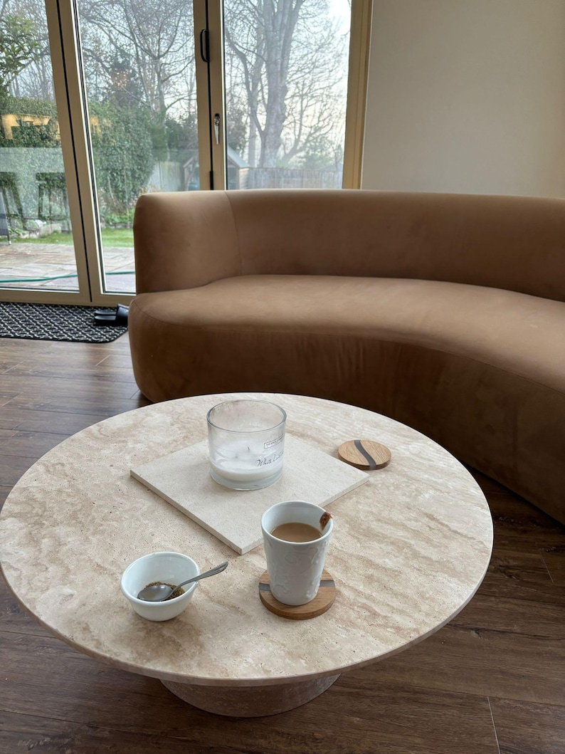 May include: A round, light-colored stone coffee table with a pedestal base. A small white bowl with a spoon, a cup of coffee, a candle in a glass jar, and two coasters are on the table. A curved brown sofa is in the background.