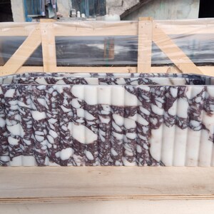 Puede incluir: Un lavabo rectangular de m&aacute;rmol con un dise&ntilde;o blanco y morado oscuro. El lavabo tiene un exterior estriado y est&aacute; colocado sobre una superficie de madera. El lavabo es un accesorio de ba&ntilde;o.