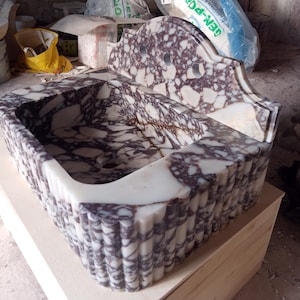 May include: A rectangular marble sink with a scalloped front edge and a decorative back. The sink features a white and purple veined pattern. The sink is made of stone and has a drain hole.