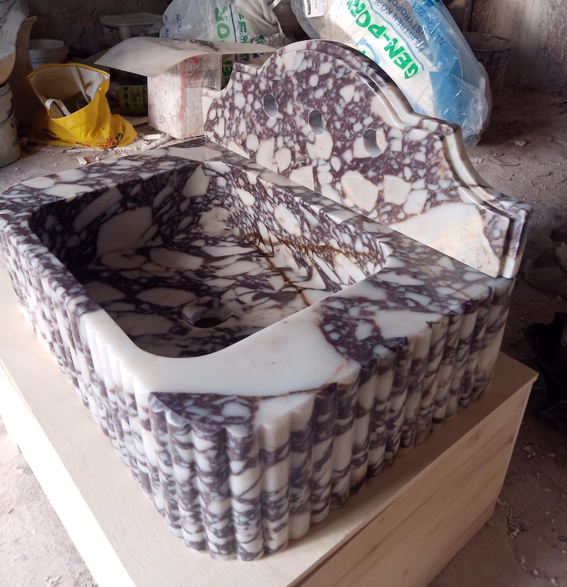May include: A rectangular marble sink with a scalloped front edge and a decorative back. The sink features a white and purple veined pattern. The sink is made of stone and has a drain hole.