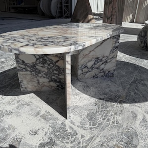 May include: A marble coffee table with a unique design. The tabletop is oval-shaped, supported by two rectangular bases. The marble features a mix of white, gray, and black veining. The table is a statement piece for any living space.