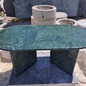 Puede incluir: Una mesa de centro ovalada de m&aacute;rmol verde con un patr&oacute;n veteado oscuro. La mesa tiene una base s&oacute;lida con dos soportes angulados. La mesa est&aacute; al aire libre, con otros elementos de piedra en el fondo.