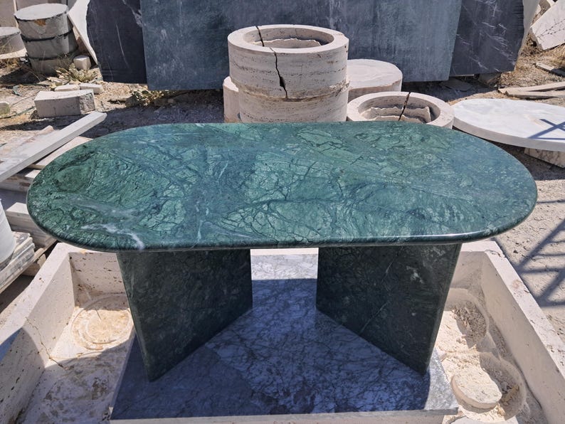 Puede incluir: Una mesa de centro ovalada de m&aacute;rmol verde con un patr&oacute;n veteado oscuro. La mesa tiene una base s&oacute;lida con dos soportes angulados. La mesa est&aacute; al aire libre, con otros elementos de piedra en el fondo.