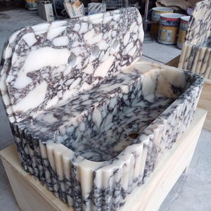 May include: A marble sink with a white and grey veined pattern. The sink has a rectangular basin and a back with three tap holes. The front and sides have a fluted design. The sink is sitting on a wooden base.