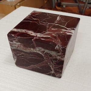 May include: A square decorative block made of dark red marble with white and light grey veining. The block has a polished finish and a geometric shape, suitable for home decor or display.