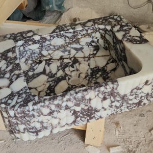 May include: A rectangular marble sink with a unique pattern of white and dark purple veining. The sink has a curved front edge and a textured, scalloped exterior. The interior features a drain hole.