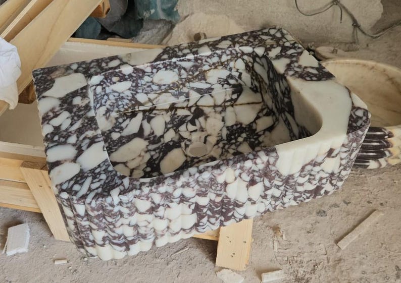 May include: A rectangular marble sink with a unique pattern of white and dark purple veining. The sink has a curved front edge and a textured, scalloped exterior. The interior features a drain hole.