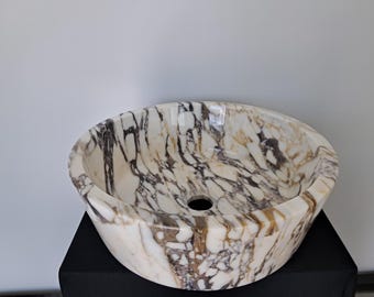 May include: Oval-shaped marble sink with a polished finish. The sink features a natural pattern of brown and black veining against a creamy white background. A central drain hole is visible. The sink is displayed on a black surface.