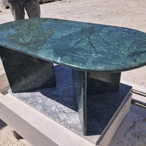 Puede incluir: Una mesa ovalada de m&aacute;rmol verde con superficie pulida. La mesa es de color verde oscuro con vetas verdes m&aacute;s claras. Se apoya en dos patas rectangulares y un soporte central. La mesa mide aproximadamente 152 cm de largo.
