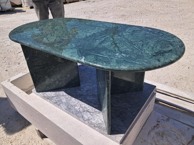 Puede incluir: Una mesa ovalada de m&aacute;rmol verde con superficie pulida. La mesa es de color verde oscuro con vetas verdes m&aacute;s claras. Se apoya en dos patas rectangulares y un soporte central. La mesa mide aproximadamente 152 cm de largo.