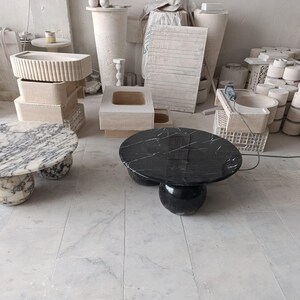 May include: Two marble coffee tables. One has a black top with white veining, the other has a white and gray marble top. Both tables have a rounded base. Various other stone objects are in the background.