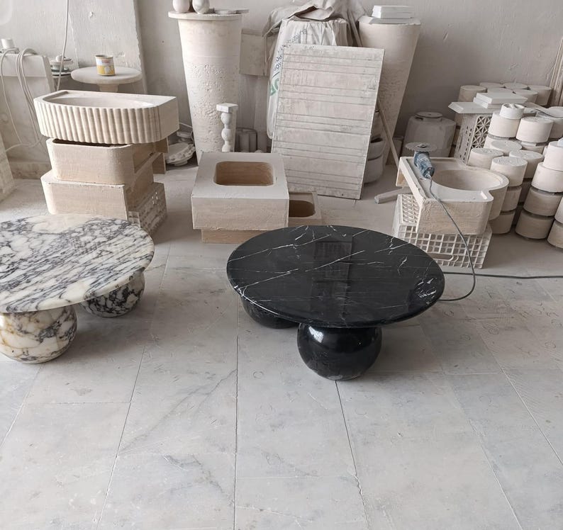 May include: Two marble coffee tables. One has a black top with white veining, the other has a white and gray marble top. Both tables have a rounded base. Various other stone objects are in the background.