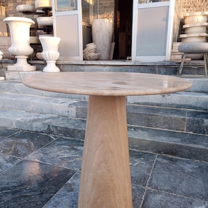 May include: A round, light brown stone table with a conical base. The table has a smooth, polished surface and stands approximately 30 inches tall. The table is made of a natural stone material, and is suitable for outdoor or indoor use.