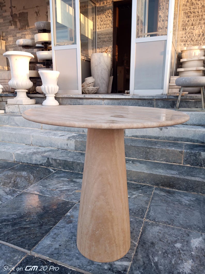 May include: A round, light brown stone table with a conical base. The table has a smooth, polished surface and stands approximately 30 inches tall. The table is made of a natural stone material, and is suitable for outdoor or indoor use.
