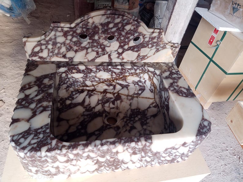 May include: A rectangular marble sink with a decorative backsplash. The sink features a white and burgundy patterned design. The backsplash has three faucet holes. The sink is made of stone and is likely for a bathroom.