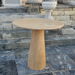 May include: A round, beige stone table with a conical base. The tabletop is approximately 30 inches in diameter. The table is made of a light-colored stone and is suitable for outdoor use.
