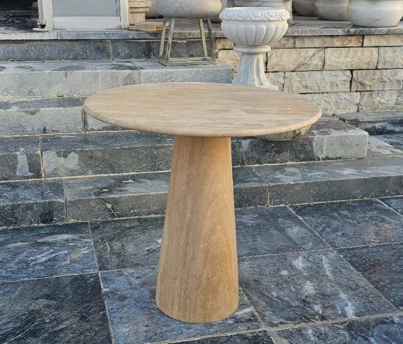 May include: A round, beige stone table with a conical base. The tabletop is approximately 30 inches in diameter. The table is made of a light-colored stone and is suitable for outdoor use.