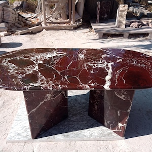 May include: Oval-shaped marble table with a rich burgundy and white veined pattern. The table rests on a matching marble base with a geometric design. A striking piece for any home.