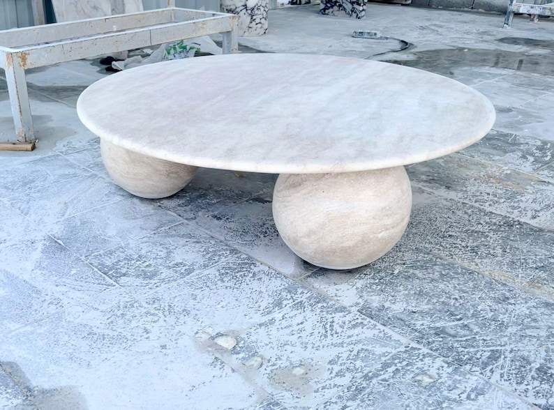 May include: A round, light-colored stone coffee table. The tabletop is supported by two spherical bases. The table has a smooth, polished surface and is likely made of marble or a similar material. The table is a neutral color and would fit in many home decor styles.
