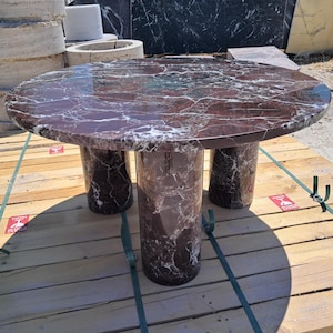 Pu&ograve; includere: Un tavolo rotondo in marmo di colore bordeaux intenso con venature bianche. Il tavolo ha tre gambe cilindriche. Il tavolo &egrave; appoggiato su un bancale di legno.