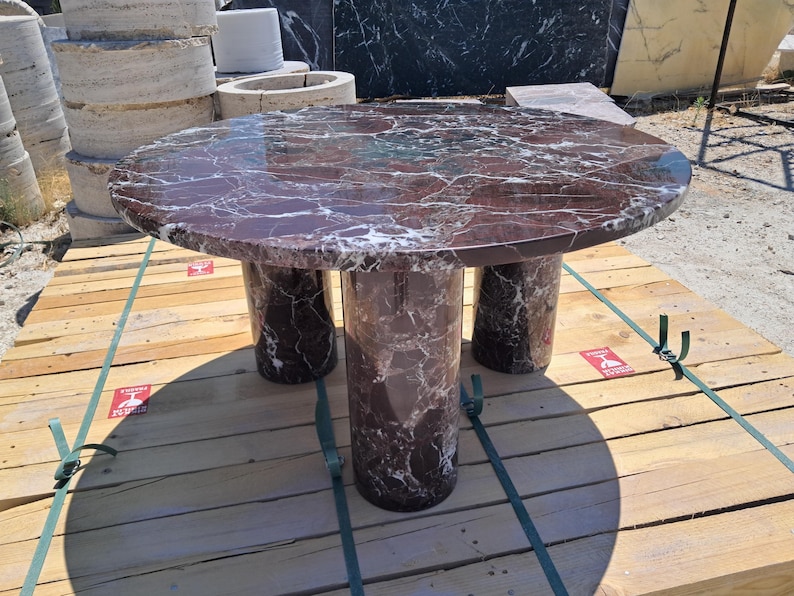Pu&ograve; includere: Un tavolo rotondo in marmo di colore bordeaux intenso con venature bianche. Il tavolo ha tre gambe cilindriche. Il tavolo &egrave; appoggiato su un bancale di legno.