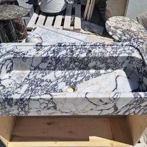 May include: A rectangular marble sink with a white base and intricate grey and black veining. The sink has a central drain and three tap holes. Other marble pieces, including a small table, are visible in the background.