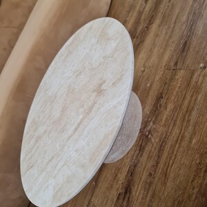 May include: Oval-shaped, light-colored stone coffee table with a smaller, circular base. The table has a natural, textured appearance and sits on a wooden floor. The table's design is simple and modern.
