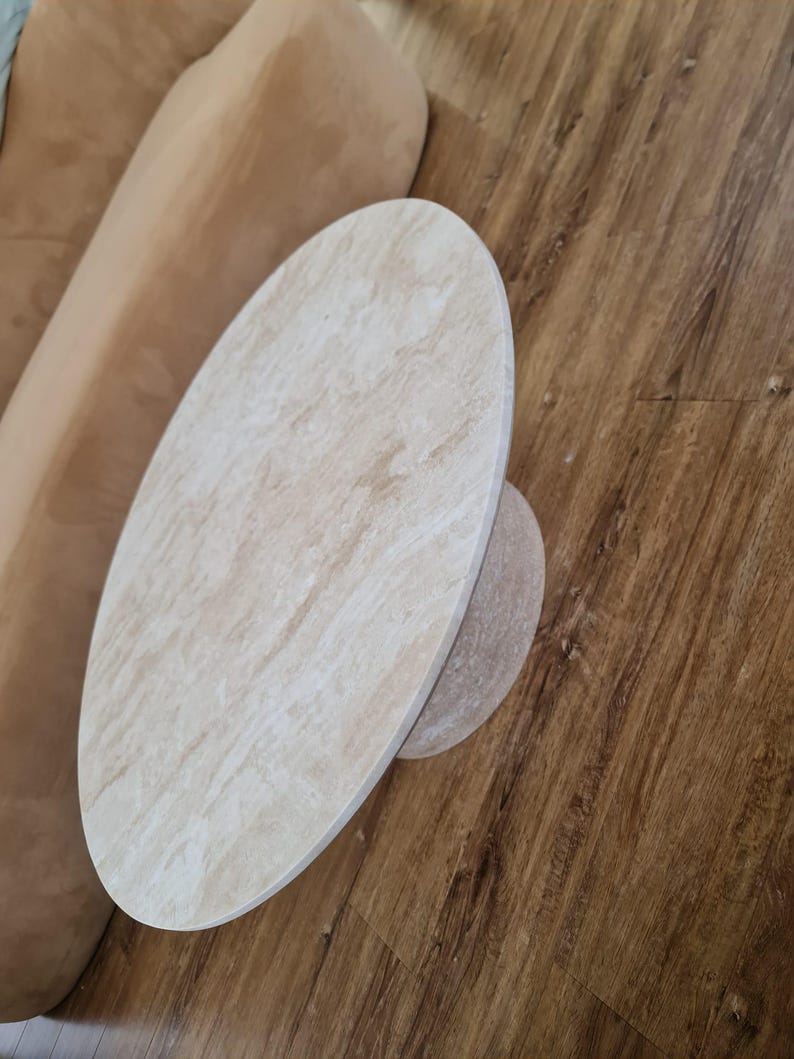 May include: Oval-shaped, light-colored stone coffee table with a smaller, circular base. The table has a natural, textured appearance and sits on a wooden floor. The table's design is simple and modern.
