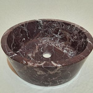 May include: A round, burgundy marble sink with white veining. The vessel sink has a smooth, polished finish and a central drain hole. The sink is a bathroom fixture.