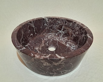 May include: A round, burgundy marble sink with white veining. The vessel sink has a smooth, polished finish and a central drain hole. The sink is a bathroom fixture.