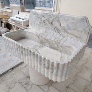 May include: A marble sink with a scalloped front edge and a backsplash featuring three tap holes. The sink is supported by a cylindrical pedestal. The marble has a white and grey veined pattern.