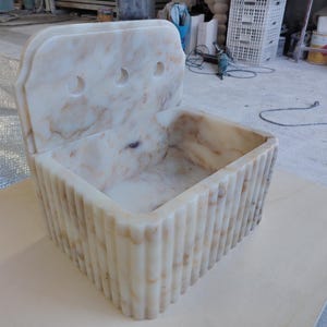 May include: A rectangular, off-white marble sink with a decorative back and three circular holes. The sink's front and sides feature vertical, ridged detailing. The marble has brown and beige veining throughout, creating a natural pattern.