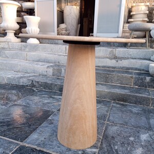 May include: A light brown stone table with a round top and a conical base. The table is made of a light brown stone material. The table is outdoors, with other stone objects in the background.