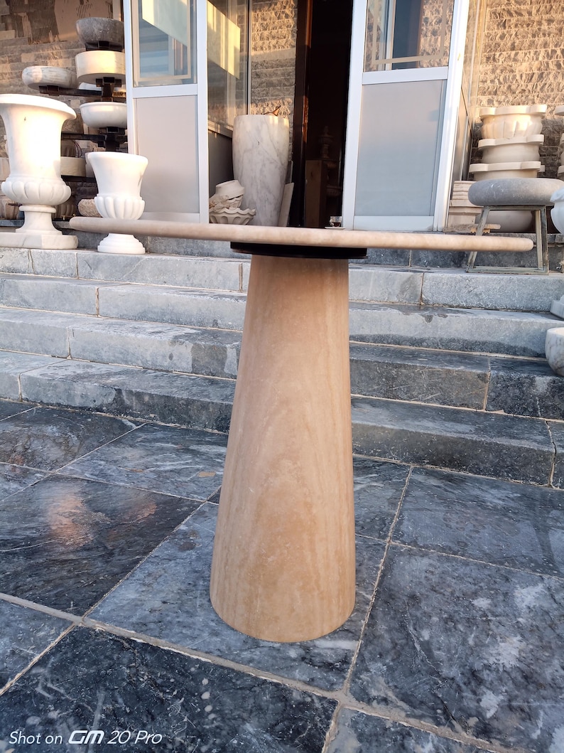 May include: A light brown stone table with a round top and a conical base. The table is made of a light brown stone material. The table is outdoors, with other stone objects in the background.