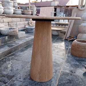 May include: A light brown stone table with a round top and a conical base. The table is approximately 36 inches in diameter and stands on a gray stone surface. Other stone objects are visible in the background.
