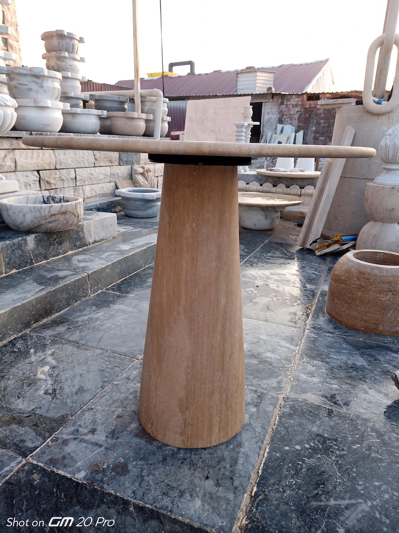 May include: A light brown stone table with a round top and a conical base. The table is approximately 36 inches in diameter and stands on a gray stone surface. Other stone objects are visible in the background.