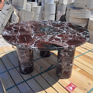 Pu&ograve; includere: Un tavolo rotondo in marmo con una superficie venata di rosso scuro e bianco. Il tavolo &egrave; sostenuto da tre gambe cilindriche in marmo abbinato. Il tavolo &egrave; su un pallet di legno.