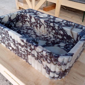 Puede incluir: Lavabo rectangular de m&aacute;rmol con un dise&ntilde;o blanco y morado. El lavabo tiene un borde festoneado y est&aacute; colocado sobre una superficie de madera clara. Un accesorio de ba&ntilde;o &uacute;nico.