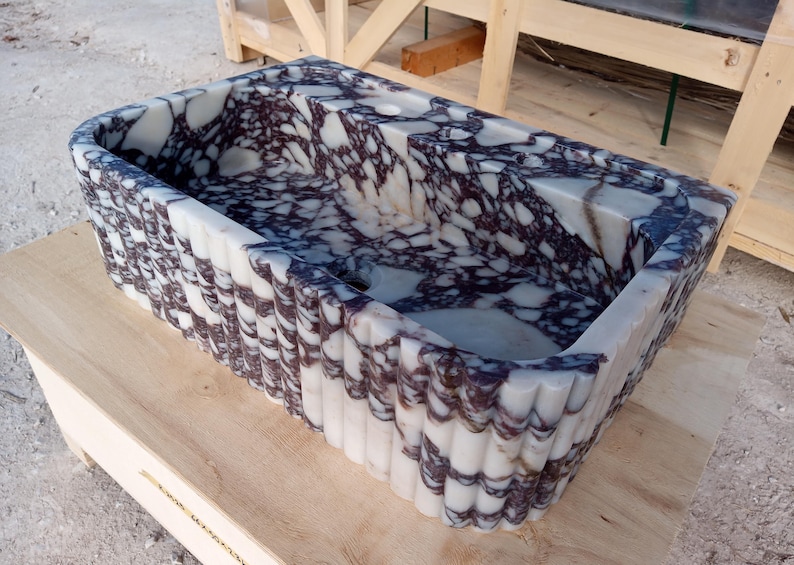 Puede incluir: Lavabo rectangular de m&aacute;rmol con un dise&ntilde;o blanco y morado. El lavabo tiene un borde festoneado y est&aacute; colocado sobre una superficie de madera clara. Un accesorio de ba&ntilde;o &uacute;nico.