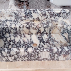 May include: A rectangular marble sink with a cream and dark gray patterned design. The sink has a built-in backsplash and a scalloped edge. It features two faucet holes and a central drain hole.
