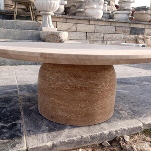 May include: A round, beige stone table with a cylindrical base. The tabletop is smooth and flat, while the base has a textured, layered appearance. The table is made of a natural stone material and is suitable for outdoor use.