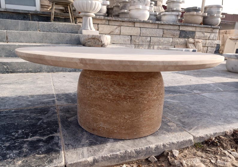 May include: A round, beige stone table with a cylindrical base. The tabletop is smooth and flat, while the base has a textured, layered appearance. The table is made of a natural stone material and is suitable for outdoor use.